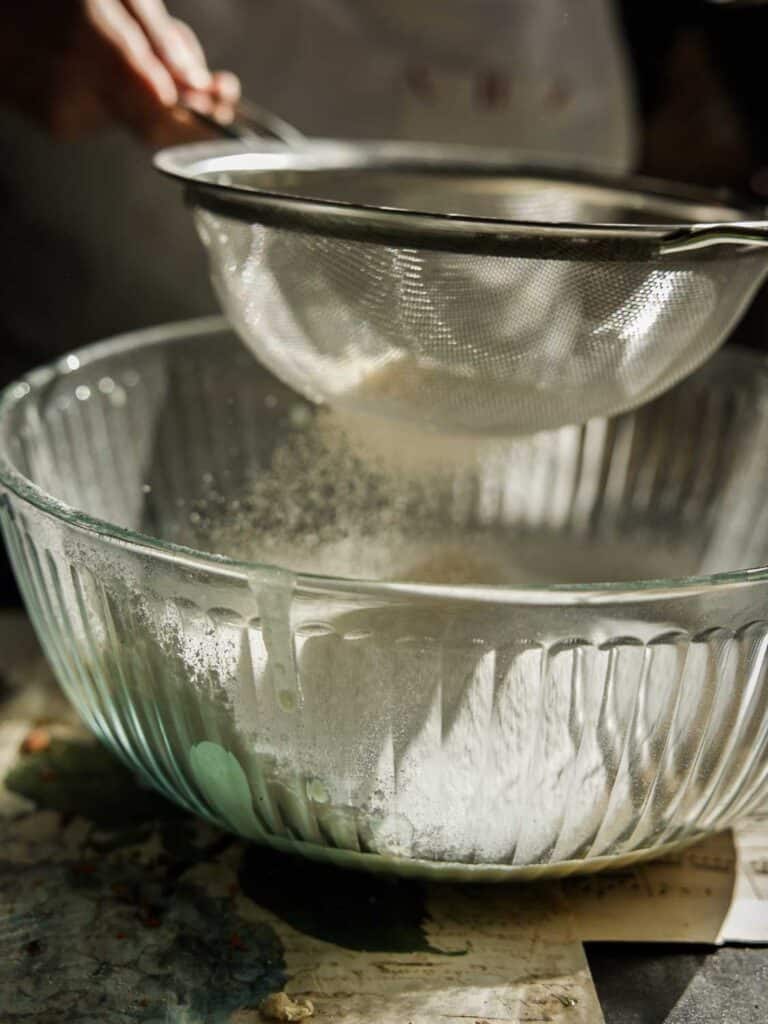 A person is sifting flour into a large glass bowl using a metal sieve. The flour is falling through the sieve, creating a light dust cloud in the bowl. The scene is warmly lit, highlighting the baking process.